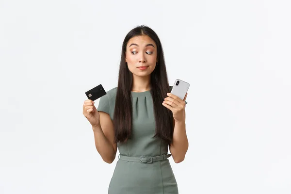 中小企業の所有者は、女性起業家の概念。躊躇懐疑的なアジアの女性は、新しい銀行アプリケーションをしようとし、携帯電話を保持しながら、クレジットカードで疑わしい見て、白い背景 — ストック写真