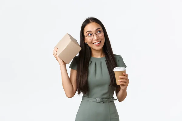 Intrigued smiling asian female drinking coffee, holding mug and shaking box as trying guess what inside, looking curious, receiving parcel from online store, standing white background — Stock Photo, Image