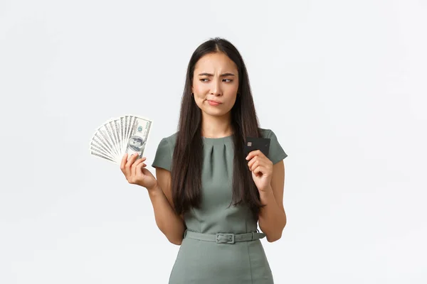 Small business owners, women entrepreneurs concept. Indecisive asian woman thinking while holding credit card and cash, perplexed how to pay for purchase, standing white background thoughtful — Stock Photo, Image