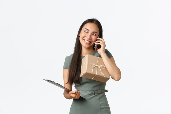 Smiling pretty asian female shop owner in dress, calling customer, holding package box for shipping to client, checking info in clipboard, making phone-call to delivery company, manage business