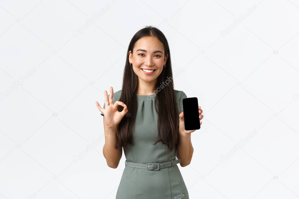 Small business owners, women entrepreneurs concept. Confident smiling asian businesswoman showing new phone application, make okay gesture and introduce mobile app on screen, white background