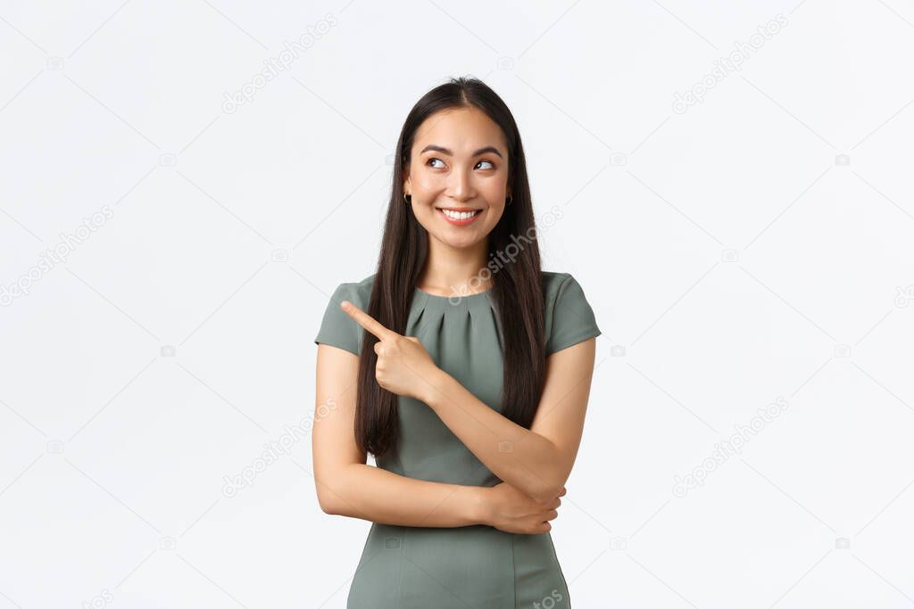 Small business owners, women entrepreneurs concept. Intrigued happy asian female in dress found excellent choice, pointing finger left and looking interested, smiling satisfied, white background