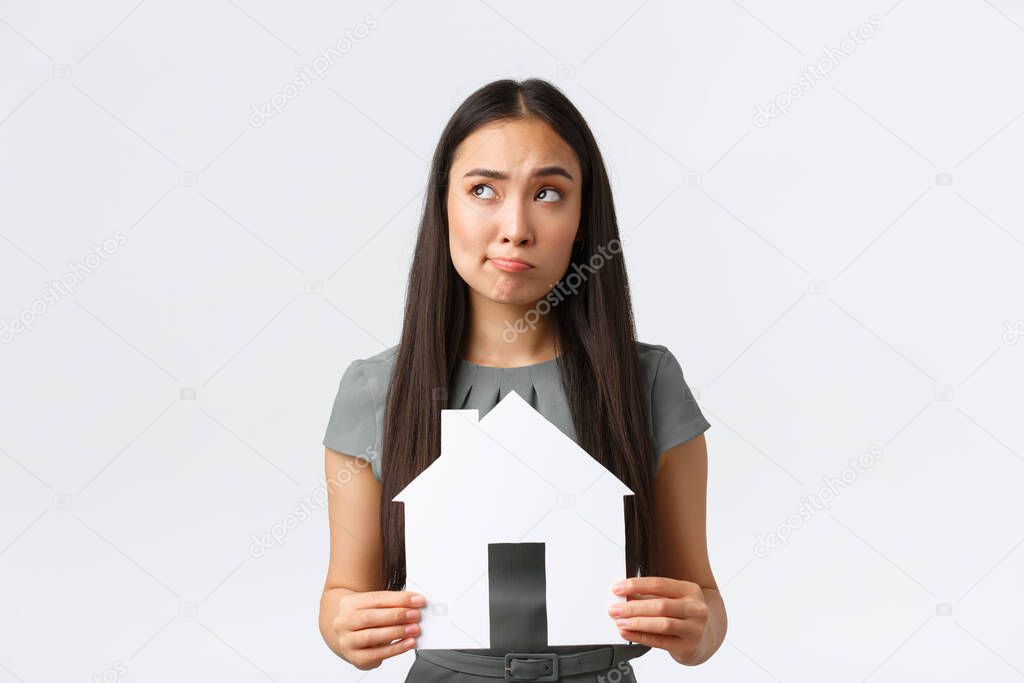 Insurance, loan, real estate and family concept. Indecisive and confused asian woman thinking about buying or renting home, holding paper house and looking away thoughtful, white background