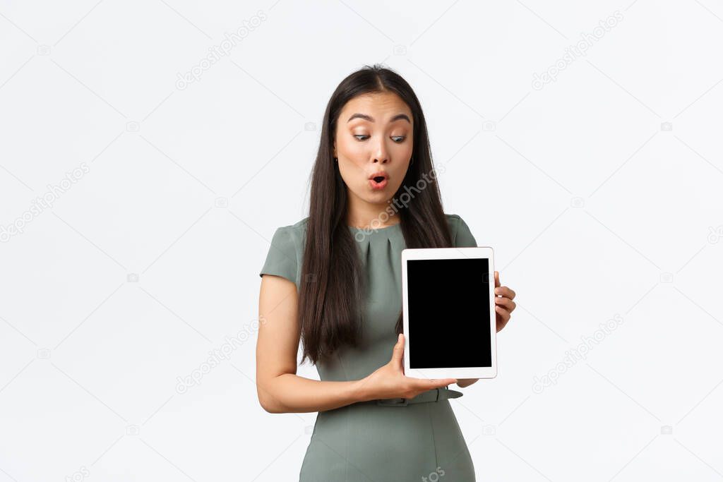 Small business owners, startup and work from home concept. Amazed and impressed asian woman in dress looking at digital tablet screen with astounded expression, checking out new items in stock