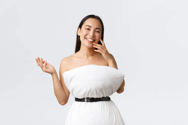 Ομορφιά, μόδα και τα μέσα κοινωνικής δικτύωσης έννοια. Αρκετά χαρούμενη Ασιάτισσα που γελάει και επιδεικνύει το νέο της ντύσιμο από μαξιλάρι και ζώνη, ποζάρει με μαξιλάρι πάνω από λευκό φόντο — Φωτογραφία Αρχείου