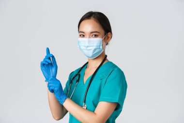 Covid-19, koronavirüs hastalığı, sağlık çalışanları konsepti. Profesyonel kendine güvenen Asyalı kadın doktor, tıbbi maske ve önlük giyen hemşire muayene için lastik eldiven takıyor, beyaz arka plan.