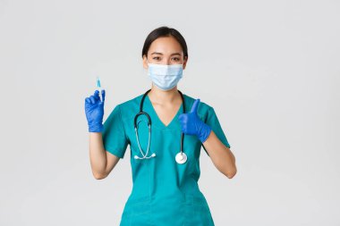 Covid-19, koronavirüs hastalığı, sağlık çalışanları konsepti. Hoş gülümseyen Asyalı kadın doktor, tıbbi maske ve eldiven giyen doktor, baş parmağını kaldır, aşı ile şırınga tut.