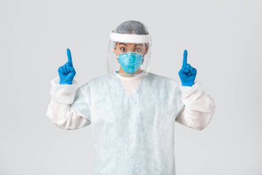 Covid-19, koronavirüs hastalığı, sağlık çalışanları konsepti. Şaşırmış ve şaşırmış Asyalı kadın doktor, solunum cihazları ve kişisel koruma ekipmanları ile reklamları işaret ediyor.