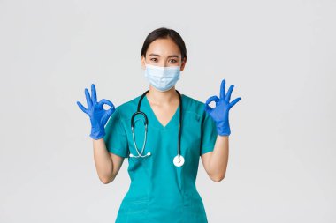 Covid-19, koronavirüs hastalığı, sağlık çalışanları konsepti. Gülümseyen profesyonel ve kendinden emin Asyalı doktor, tıbbi maske, eldiven ve önlük giyen doktor iyi niyet göstergesi, kaliteli hizmet garantisi