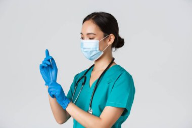 Covid-19, koronavirüs hastalığı, sağlık çalışanları konsepti. Profesyonel gülümseyen Asyalı hemşire, ameliyat önlüğü ve tıbbi maske giyen doktor muayene için lastik eldiven taktı, hasta muayene etti.