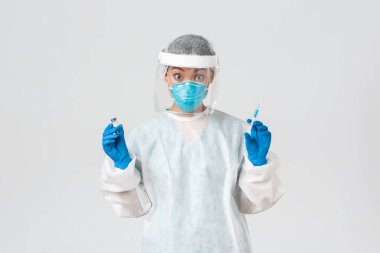 Covid-19, koronavirüs hastalığı, sağlık çalışanları konsepti. Şaşırmış ve heyecanlı Asyalı doktor, kişisel koruyucu ekipman enfeksiyonisti, elinde şırınga ve aşı ampulü var.