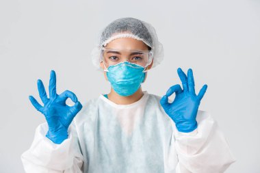 Covid-19, koronavirüs hastalığı, sağlık çalışanları konsepti. Kişisel koruma ekipmanlarıyla memnun olmuş, ciddi görünümlü Asyalı bir kadının yakın plan çekimi tamam bir jest yap, onayla ya da garanti et