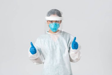 Covid-19, koronavirüs hastalığı, sağlık çalışanları konsepti. Ciddi görünüşlü, kendine güvenen genç Asyalı doktor, kişisel koruma ekipmanlarında enfeksiyoncu baş parmağını kaldırarak tehlikeli bölgeye giriyor.