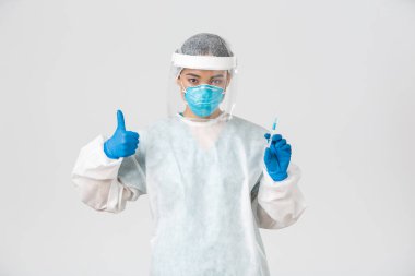 Covid-19, koronavirüs hastalığı, sağlık çalışanları konsepti. Kişisel koruma ekipmanlarında kendine güvenen ciddi Asyalı kadın doktor, baş parmağını kaldır ve aşı ile şırıngayı tut, beyaz arka plan