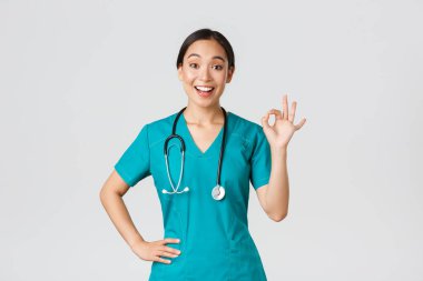 Covid-19, sağlık çalışanları, salgın konsepti. Şaşırmış ve mutlu Asyalı kadın doktor, önlüklü hemşire iyi bir jest yapıyor ve şaşırmış bir şekilde gülümsüyor, iyi iş çıkarıyor, birisiyle aynı fikirde.
