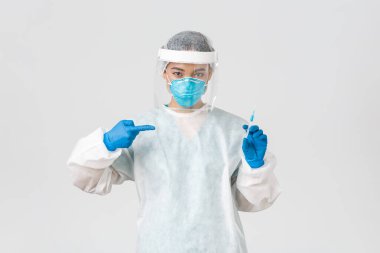 Covid-19, koronavirüs hastalığı, sağlık çalışanları konsepti. Ciddi bir Asyalı kadın doktor, PPE kişisel koruma ekipmanı doktoru, aşı şırıngasını işaret eden, beyaz arka plan