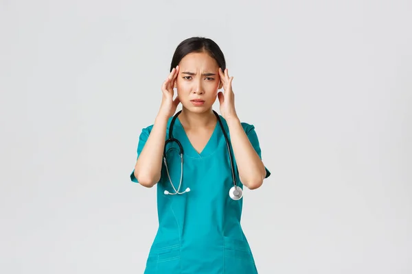 Covid-19, egészségügyi dolgozók, világjárvány. Túlhajszolt és kimerült ázsiai női orvos, hányinger, köpeny viselése, fejérintés, fejfájás vagy magas láz panasza — Stock Fotó