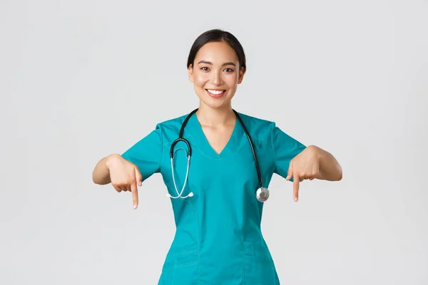 Covid-19, healthcare workers, pandemic concept. Smiling pleasant asian female doctor, therapist or physician in scrubs with stethoscope, pointing fingers down, show clinic banner — Stock Photo, Image