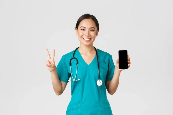 Covid-19, trabajadores de la salud y concepto de medicina en línea. Sonriente mujer asiática confiada médico, médico en matorrales mostrar signo de paz y pantalla móvil, promover la aplicación de consulta, fondo blanco — Foto de Stock