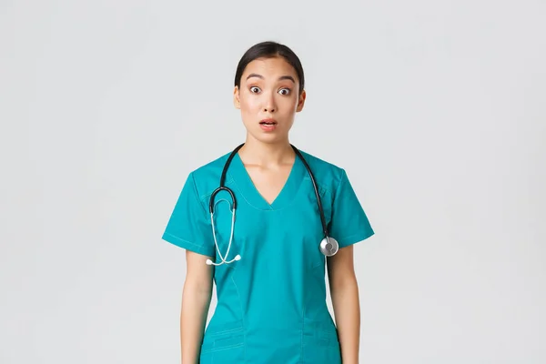 Covid-19, healthcare workers, pandemic concept. Impressed and surprised asian nurse in scrubs drop jaw and stare at camera speechless, looking amazed over white background