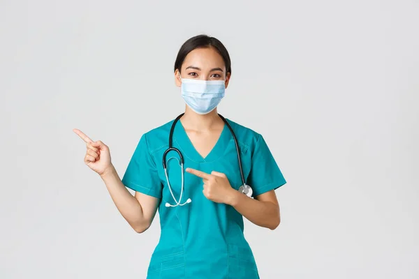 Covid-19, choroba koronawirusowa, koncepcja pracowników służby zdrowia. Profesjonalny pewny azjatycki lekarz, lekarz w masce medycznej i peelingi wskazując palcem w lewo, pokazując reklamy — Zdjęcie stockowe