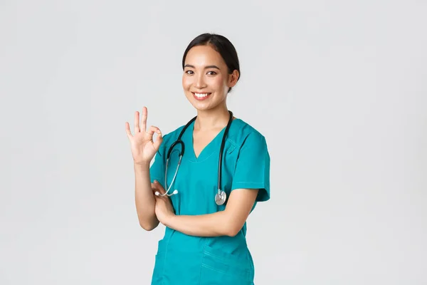 Covid-19, healthcare workers, pandemic concept. Confident professional asian doctor, female physician in scrubs, showing okay gesture, guarantee quality of services in hospital, approve and agree — Stock Photo, Image