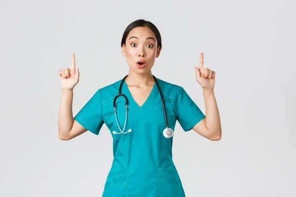 Covid-19, 의료 종사자, 전염병같은 개념. 당황하고 흥분 한 동양 여성 간호사, 수술복을 입고 흥미 로운 프로 모를 묻는 여성 의사, 손가락을 치켜들고 진료소 광고를 보여 주는 모습 — 스톡 사진