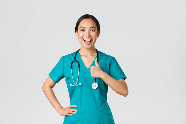 Ковід-19, медичні працівники, пандемія. Усміхнений біт, впевнена жінка-азіатка медсестра в скрабах все під контролем, показати великі пальці в погодженні, гарантувати якість обслуговування в клініці — стокове фото