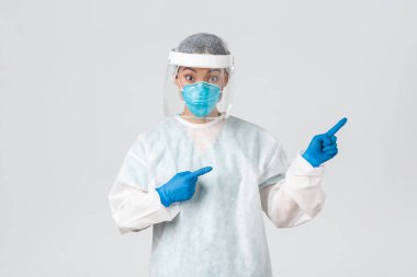 Covid-19, koronavirüs hastalığı, sağlık çalışanları konsepti. Şaşırmış ve şaşırmış Asyalı kadın doktor, kişisel koruma ekipmanları içinde teknoloji laboratuarı çalışanı şaşırmış ve parmağıyla işaret ediyor.