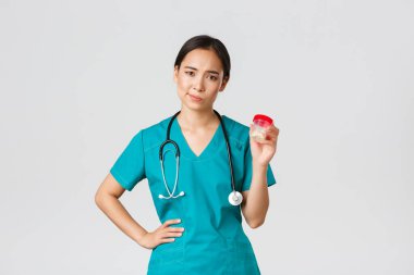 Sağlık çalışanları, virüsü önleme, karantina kampanyası konsepti. Şüpheci ve hoşnutsuz Asyalı kadın doktor, hemşire ilaç önermiyor, isteksiz sırıtıyor ve hapları göstermiyor, beyaz bir geçmişi var.