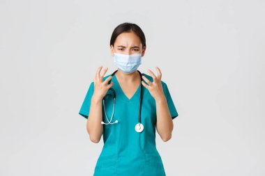 Covid-19, coronavirus disease, healthcare workers concept. Angry and pissed-off asian female doctor, physician in medical mask and scrubs, clench fists outraged, looking furious, white background
