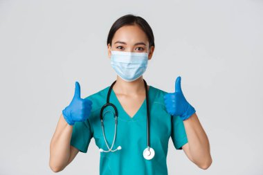 Covid-19, koronavirüs hastalığı, sağlık çalışanları konsepti. Kendinden emin gülümseyen Asyalı doktor, tıbbi maske takan hemşire ve lastik eldiven, onaylama için onay ver, hizmet et, bilgi topla.
