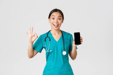Covid-19, sağlık çalışanları ve online tıp konsepti. Koronavirüs sırasında yeni bir online danışma yöntemi deneyen Asyalı kadın doktor akıllı telefon uygulaması ve tamam jest gösteriyor.