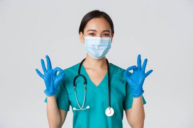 Covid-19, koronavirüs hastalığı, sağlık çalışanları konsepti. Kendinden emin gülümseyen Asyalı kadın doktor, tıbbi maske ve eldivenli hemşire, onaylı, beyaz arka planda iyi niyet göstergesi göster.
