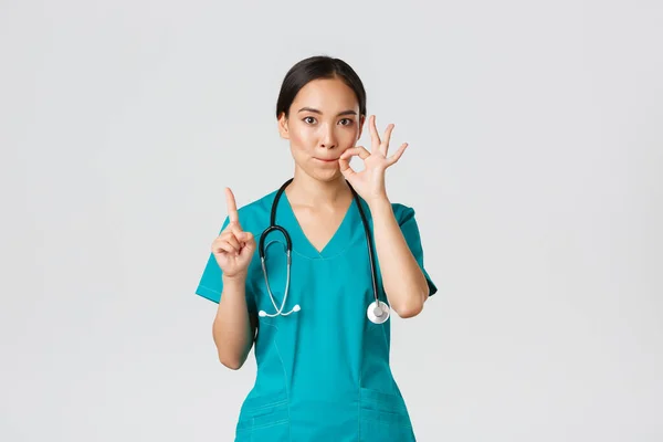 Covid-19, healthcare workers, pandemic concept. Serious-looking worried asian female nurse, physician asking keep secret, shaking finger and showing mouth seal, zipping lips gesture