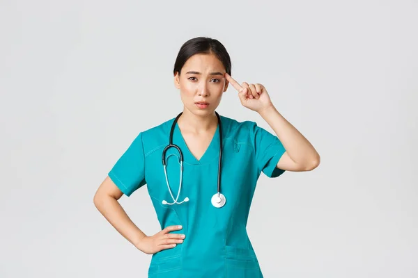 Covid-19, zdravotníci, pandemický koncept. Otravný skeptický asijské žena zdravotní sestra nebo lékař pokárat někoho hrát šílený nebo hloupý, roll prst přes spánek s odsuzující tvář — Stock fotografie