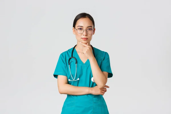 Covid-19, pracownicy służby zdrowia, koncepcja pandemii. Poważnie wyglądająca młoda kobieta internet, azjatycka pielęgniarka lub lekarz w fartuchu i okularach myśląc, rozważając lub dokonując wyboru, zbadać pacjenta — Zdjęcie stockowe