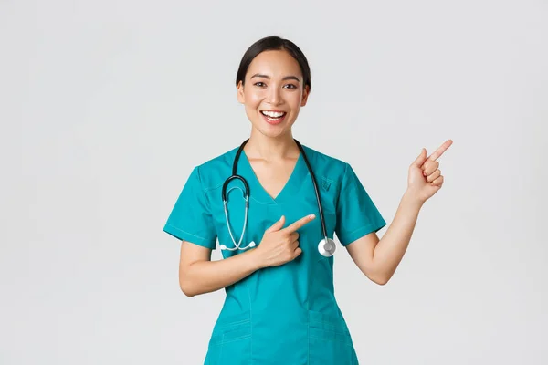 Covid-19, profissionais de saúde, conceito pandémico. Sorrindo alegre asiático feminino médico, terapeuta em esfrega apontando dedos canto superior direito, convidando para check-up, recomendo vacinar — Fotografia de Stock