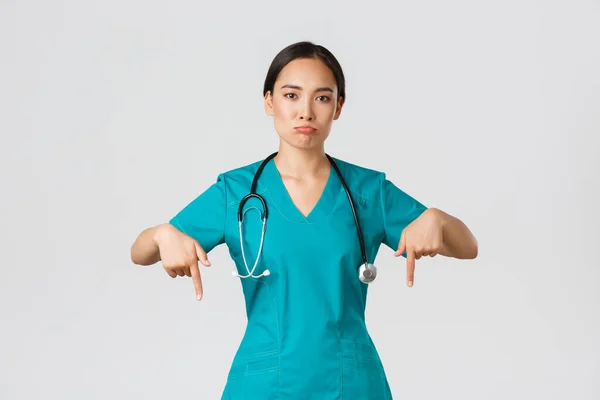 Covid-19, pracownicy służby zdrowia, koncepcja pandemii. Zdenerwowana i rozczarowana dąsająca się azjatycka terapeutka, pielęgniarka w fartuchu wskazująca palcem w dół, dąsająca się smutna, pokazująca coś z żalem — Zdjęcie stockowe