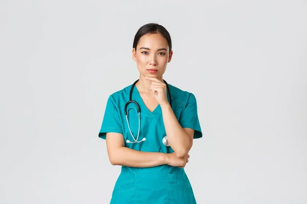 Covid-19, egészségügyi dolgozók, világjárvány. Átgondolt női orvos, ázsiai ápolónő köpenyben kíváncsi a kamera, gondolkodás, keresi a megoldást, álló fehér háttér töprengés — Stock Fotó