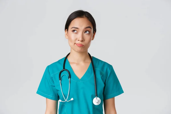 Pracownicy służby zdrowia, zapobieganie wirusom, koncepcja kampanii kwarantanny. Przemyślany azjatycki lekarz, pielęgniarka w fartuchu patrząc w lewy górny róg i uśmiech niepewny, myślenie, białe tło — Zdjęcie stockowe