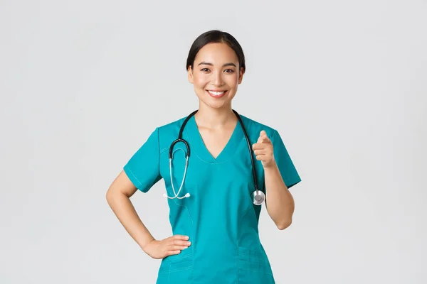 Covid-19, profissionais de saúde, conceito pandémico. Confiante sorrindo asiático profissional médico, enfermeira em esfrega apontando o dedo para a câmera, convidando para check-up ou vacinação, dar recomendação — Fotografia de Stock