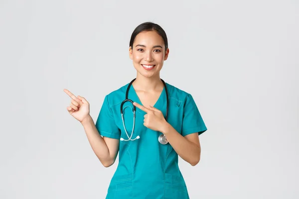 Covid-19, healthcare workers, pandemic concept. Smiling pretty asian doctor, nurse in scrubs and stethoscope, pointing fingers upper left corner, showing online clinic banner, white background — Stock Photo, Image