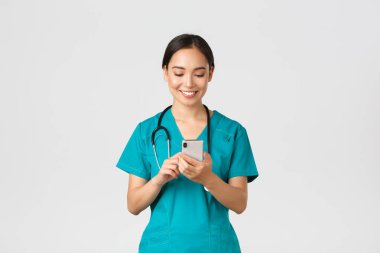 Covid-19, sağlık çalışanları ve virüs konseptini önlemek. Gülümseyen mutlu Asyalı kadın stajyer, doktor telefon görüşmesi yapıyor, cep telefonu ekranına bakıyor memnun, mesajlaşıyor ya da uygulama kullanıyor.