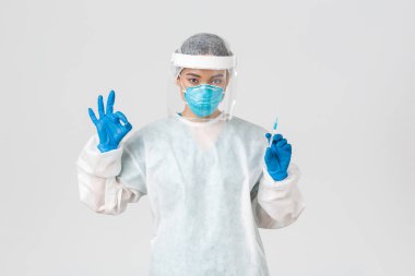 Covid-19, koronavirüs hastalığı, sağlık çalışanları konsepti. Kişisel koruma ekipmanlarında kendine güvenen ciddi Asyalı kadın doktor, iyi bir jest göster, aşıyla şırıngayı tut, beyaz arka plan