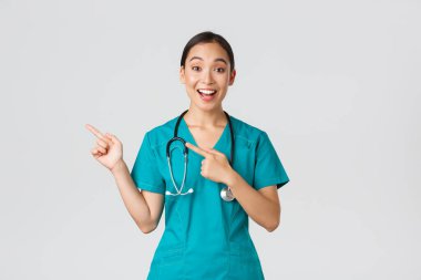 Covid-19, sağlık çalışanları, salgın konsepti. Heyecanlı Asyalı hemşire, önlüklü ve steteskoplu doktor, parmaklarını sol üst köşeyi işaret ediyor. Hekim gülümseyerek müthiş bir duyuru yapıyor.