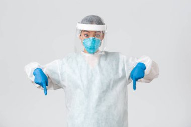 Covid-19, koronavirüs hastalığı, sağlık çalışanları konsepti. Şaşırmış ve heyecanlı kadın doktor, kişisel koruyucu ekipman hekimi parmakları aşağıda, beyaz arka planda duruyor.