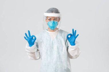 Covid-19, koronavirüs hastalığı, sağlık çalışanları konsepti. Kendinden emin ve ciddi Asyalı kadın doktor, kişisel koruma ekipmanlarında laboratuvar teknisyeni onaylı jest gösteriyor.