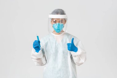 Covid-19, koronavirüs hastalığı, sağlık çalışanları konsepti. Ciddi ciddi Asyalı kadın doktor, kişisel koruma ekipmanlarında teknoloji laboratuarı çalışanı baş parmağını kaldır ve aşı ile test tüpünü tut.