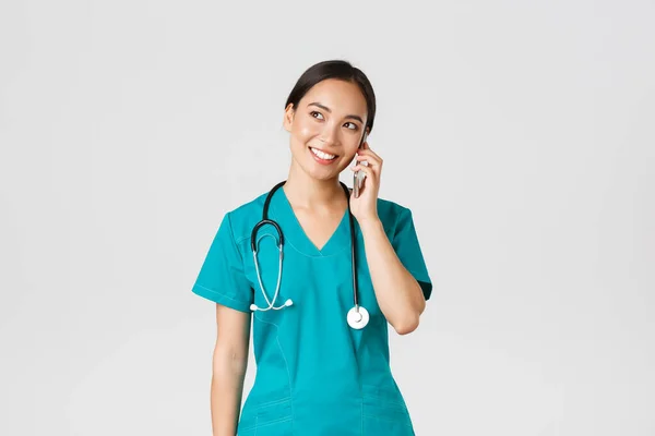 Covid-19, pracownicy służby zdrowia i zapobieganie koncepcji wirusa. Śliczny uśmiech azjatycki lekarz, lekarz w fartuchu prowadzenia rozmowy, rozmowy przez telefon i patrząc w lewym górnym rogu marzycielski — Zdjęcie stockowe